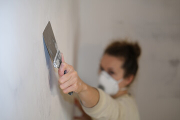 A woman worker in a respirator levels the walls with a putty knife