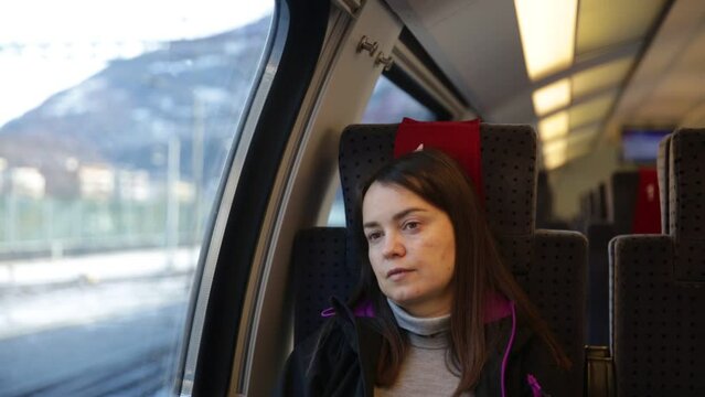Dreamy relaxed female traveler sitting near window on comfortable chair in express train, looking at picturesque mountain landscapes of Switzerland on sunny winter day behind glass. 