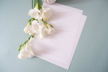 A blank letter paper with white freesia flowers and vase on white pale green background. Mother’s Day, Women7s day, Father’s Day, weeding celebration background.