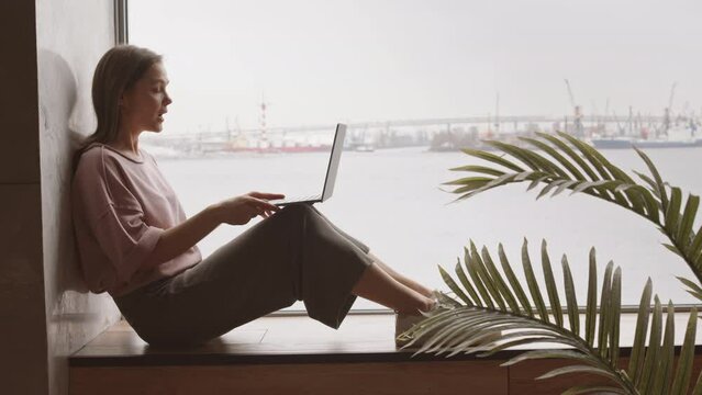 Side-view slowmo shot of young smiling woman video conferencing on laptop sitting on windowsill by panoramic window with sea view