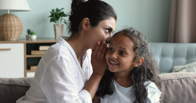 Close up Indian mother her little curious daughter share secrets seated together on sofa at home, cover mouth with palms gossiping, have fun, enjoy communication and trustworthy relationships concept