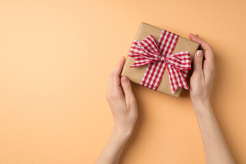 First person top view photo of st valentine's day decorations woman's hands demonstrating craft paper giftbox with checkered ribbon bow on isolated beige background with copyspace