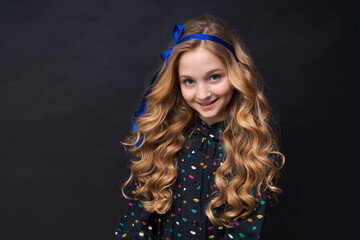 Smiling pretty little caucasian girl 12-10 posing on black background, childrens studio portrait, beautiful long wavy hair with a blue ribbon on her head. Childhood lifestyle concept