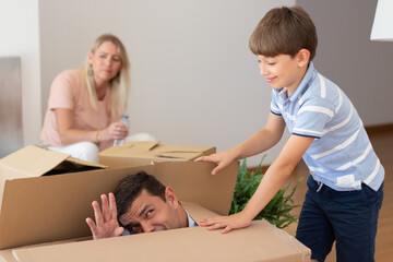 Amusing little boy with father playing in new house. Schoolboy with dark hair hiding dad in big cardboard box. Real estate, purchase, family concept
