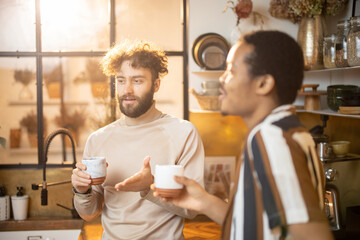 Two guys of different ethnicity having warm conversation while drinking coffee on kitchen at home. Concept of close male friendship or relationship as gay couple. Caucasian and hispanic man together