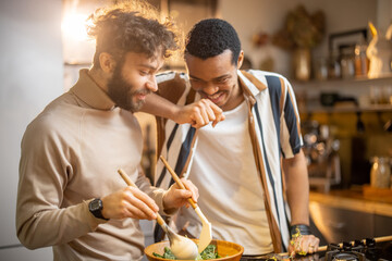 Two guys of different ethnicity having fun while making salad together on kitchen. Concept of gay couples and everyday life at home . Caucasian and hispanic man cooking healthy food - obrazy, fototapety, plakaty