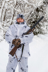 Portrait of a male hunter in a winter forest with a gun in a camouflage white suit. The concept of winter hunting.