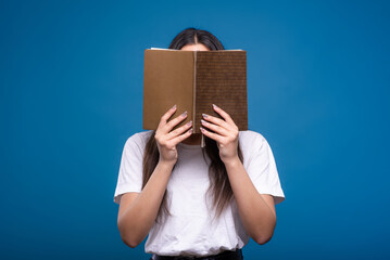 Attractive brunette girl in a white t-shirt holding a book and reading isolated on a blue studio...