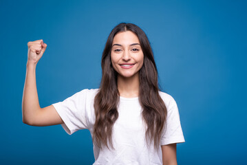 Attractive caucasian or arab brunette girl in white t-shirt clenching fist showing biceps isolated on blue studio background.