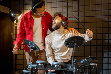 Two male close friends hug while playing electric drums, composing electronic music at small...
