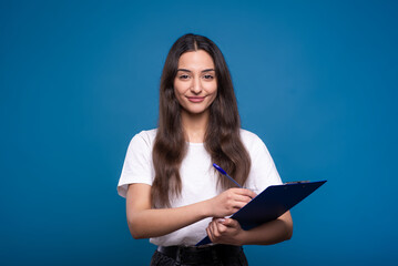Attractive caucasian or arab brunette girl in a white t-shirt holding a tablet, writing and smiling isolated on a blue studio background.