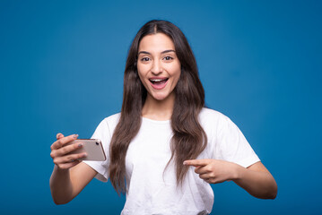 Attractive caucasian or arab brunette girl in a white t-shirt is shocked while playing on a mobile phone and pointing her finger at a smartphone isolated on a blue studio background.
