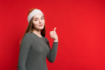 A beautiful smiling girl in a new year's hat shows her thumbs up to the camera. Thumbs up gesture. Camera. New year. Christmas
