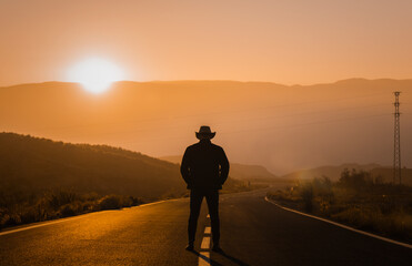 Fototapeta na wymiar Silhouette of adult man in cowboy hat standing on country road during sunset. Almeria, Spain