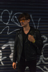 Portrait of young stylish brunet caucasian man in black leather jacket and yellow sunglasses. Standing near the wall with graffiti. High quality photo