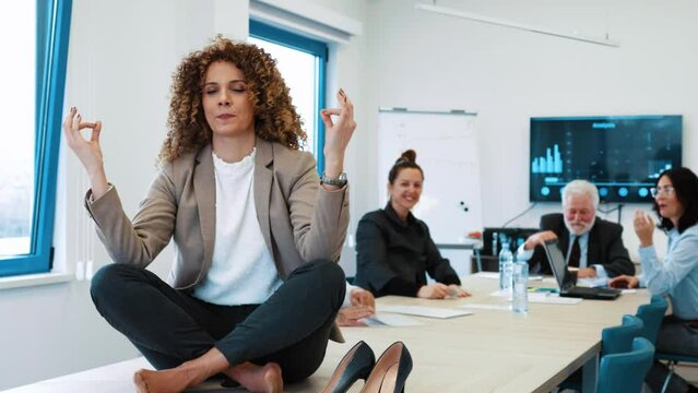Young business girl trying to meditate in lotus position sitting on table in office while her colleagues bother her and make noise  