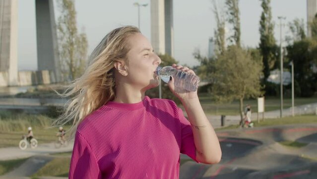 Beautiful girl drinking water after skateboarding in park. Cute Caucasian girl choking on water, having rest after training in skate park, spending time outdoor on sunset. Sport, relaxation concept