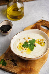 Hummus dip with chickpea and parsley in ceramic bowl on concrete surface with toasted baguette slices and olive oil on wooden board