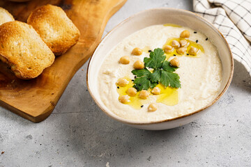 Hummus dip with chickpea and parsley in ceramic bowl on concrete surface with toasted baguette slices and olive oil on wooden board