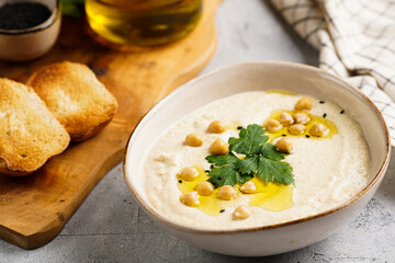 Hummus dip with chickpea and parsley in ceramic bowl on concrete surface with toasted baguette slices and olive oil on wooden board