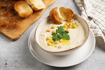 Hummus dip with chickpea and parsley in ceramic bowl on concrete surface with toasted baguette slices and olive oil on wooden board