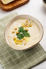Hummus dip with chickpea and parsley in ceramic bowl on marble surface with toasted baguette slices and olive oil on wooden board