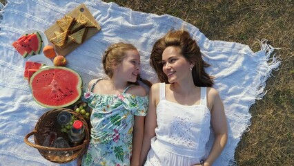 Happy mother touches nose of little daughter lying on white blanket with delicious fruits prepared for picnic on sunny summer day top view - Powered by Adobe