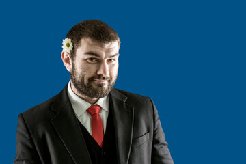 A handsome bearded brunette in a suit with a flower behind his ear looks cutely into the camera. A romantic person
