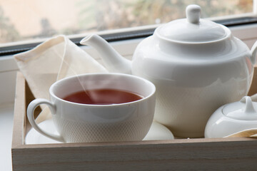 A white cup with hot tea on a saucer, a teapot, a sugar bowl and a cloth napkin on a wooden tray against the background of the window. Close-up.