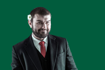 A handsome bearded brunette in a suit with a flower behind his ear looks cutely into the camera. A romantic person. Gentleman.