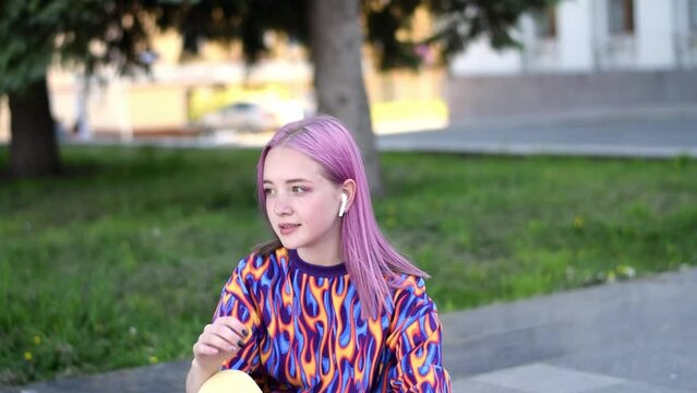 Smiling pink-haired teenage hipster girl in colorful T-shirt and earphones is  sitting on a skateboard outside on a summer day.Generation Z style.Slow motion.