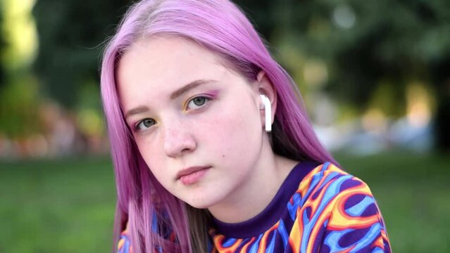 Pink-haired teenage hipster girl in a colorful bright T-shirt and earphones is using a smartphone on a city street on a summer day.Summer concept.Generation Z style.Social media concept.Slow motion.