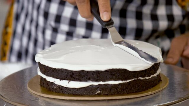 An Indian cook wearing a checked apron spreading fresh cream on a chocolate cake. Confectioner decorating a birthday cake using a spatula at a commercial bakery - culinary design  cooking ingredient