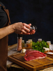 The chef sprinkles spices on a piece of raw meat - pork, beef, lamb on a wooden cutting board. Background - meat with vegetables on a black background. The concept is a recipe for cooking meat.