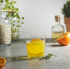Homemade orange lemonade with thyme at grey concrete kitchen table with orange half, glass jar with oats and bottle. Preparing refreshing summer drink at home. Front view.