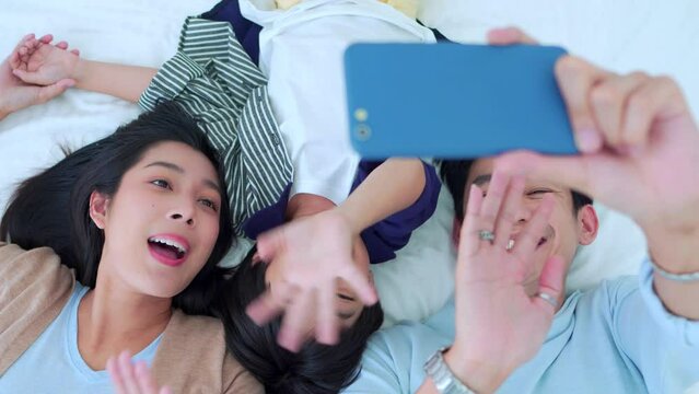 Happy family time, Taking selfie mobile phone of Asian parents dad, mom and son laying together on bed in bedroom with happy, laughing smiles. Weekend parenting with love good time relaxing at home.
