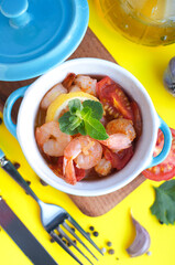 Baked shrimps with tomatoes, garlic, lemon, olive oil,  mint and fork and knife in blue bowls on a yellow background. Baked shrimps on wooden board. Top view