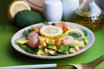 Salad with shrimps, avocado, corn and lemon with olive oil on a green background. Salad with shrimps and avocado on white plate with salt shaker, fork and knife
