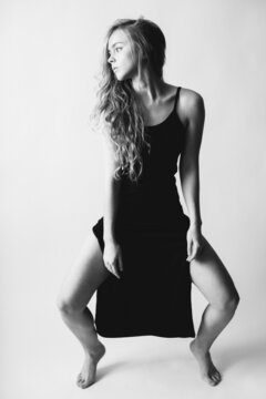 Beautiful girl with long hair in a black nightgown dress with straps spontaneously moves and poses, a dancing girl with a good mood. Black and white portrait of a young girl on a white background