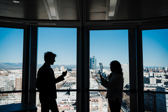 Silhouette of businessman and businesswoman using smart phones by glass wall in office - Powered by Adobe