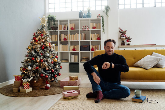 Happy man checking smart watch sitting by Christmas tree at home