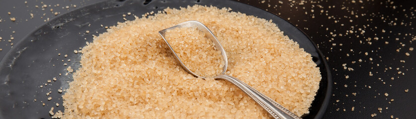 A fragment of a glass black plate filled with brown sugar. There is sugar scattered around and a spoon lying on the plate.