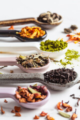 Various types of dry tea in spoons on a white marble background. Green, matcha, citrus, black and herbal detox tea.