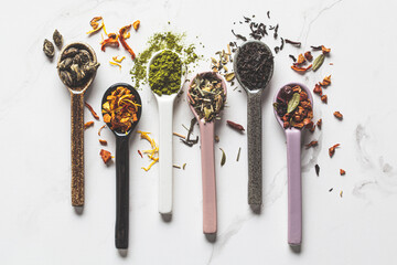 Various types of dry tea in spoons on a white marble background, top view. Green, matcha, citrus,...