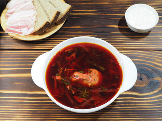 Delicious hot beetroot soup in a white bowl, sour cream, rye bread with bacon on a wooden table, top view, flat layout. Traditional red borscht with meat and vegetables