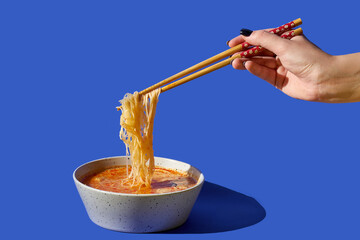 Woman's hand with chopsticks takes noodles out of ramen soup in a bowl against a blue background