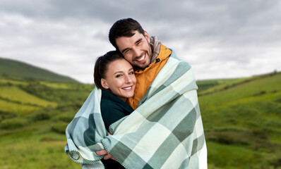 love, travel and tourism concept - happy smiling couple in warm blanket over farmland fields and hills at wild atlantic way in ireland background