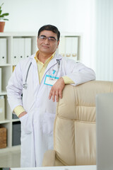 Portrait of confident smiling chief physician standing at his chair in medical office