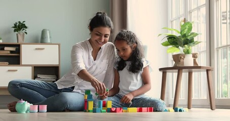 Loving Indian mom spend time with her cute little daughter settled down on warm floor in living room, play together multi colored wooden cubes. Child development and growth, fun, games at home concept