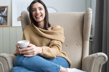 Positive young lady in casual wear sitting in armchair, warming hands on cup of hot coffee,...